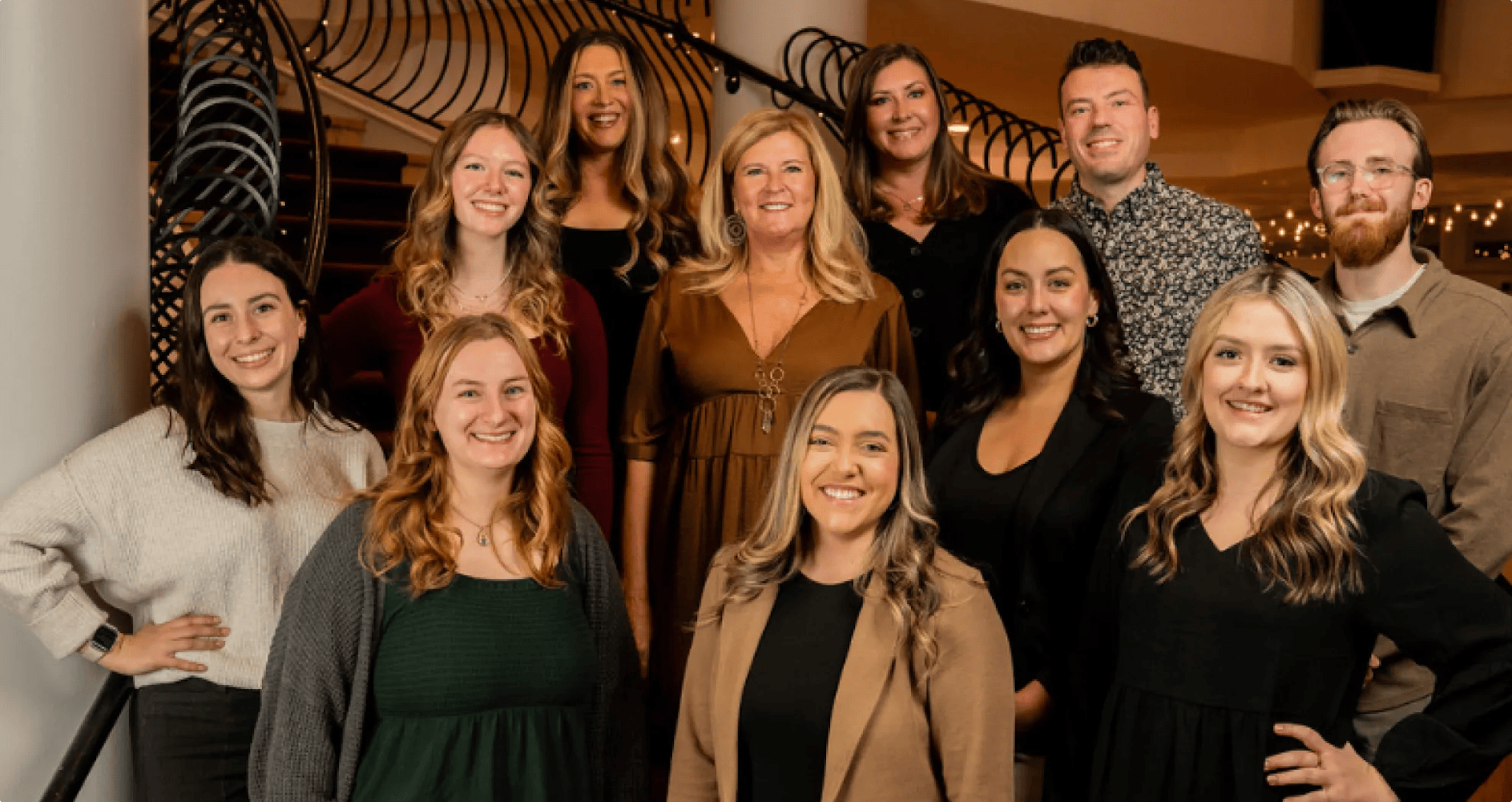 Group of eleven smiling adults posing indoors with a curved staircase and string lights in the background.